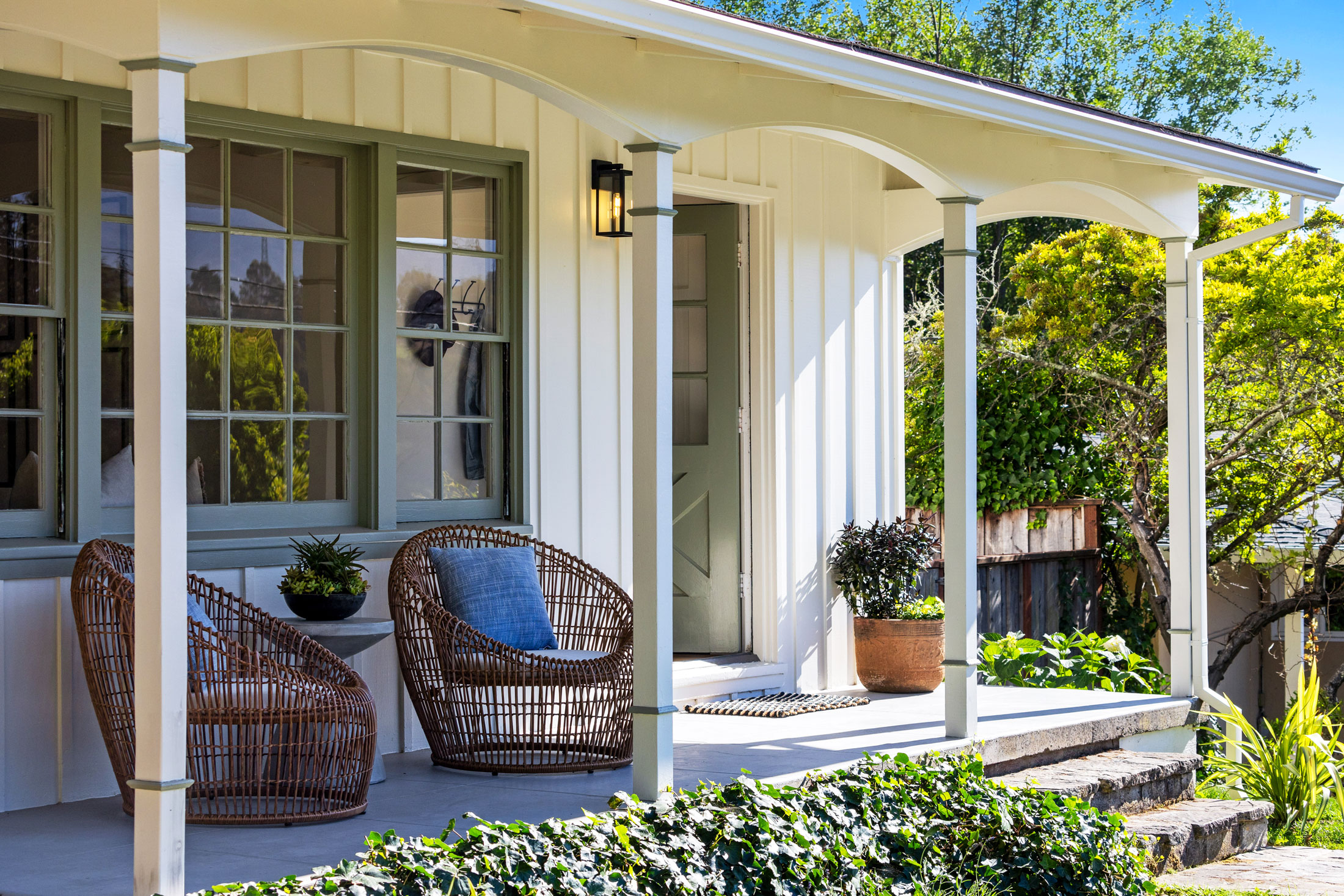 114 Richardson Drive A cozy front porch at 114 Richardson Drive Mill Valley features two wicker chairs, a small table with potted plants, a white exterior, green-trimmed windows, and lush greenery surrounding the steps and garden area.