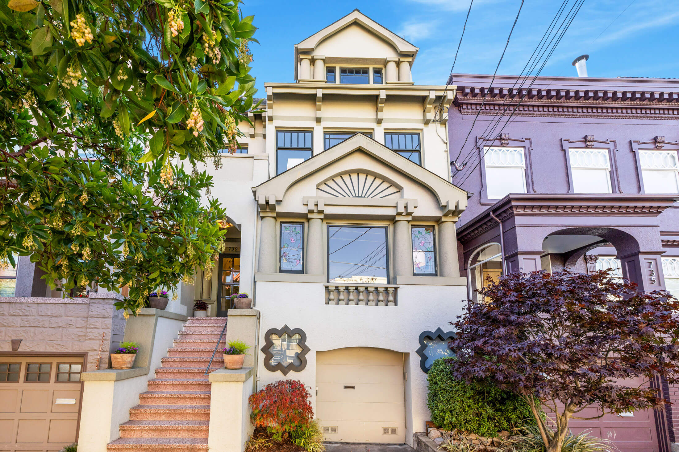 A two-story house at 739 10th Avenue, San Francisco, features large windows, decorative architectural details, a small garage, and a staircase leading to the entrance. Surrounded by trees and plants, it sits beside a purple neighboring building.