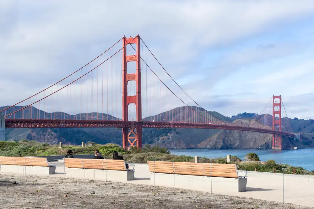 Tunnel Tops view of Golden Gate Bridge from Presidio The Golden Gate Bridge spans across the water with hills in the background; several empty benches and a few people are visible in the foreground under a partly cloudy sky.