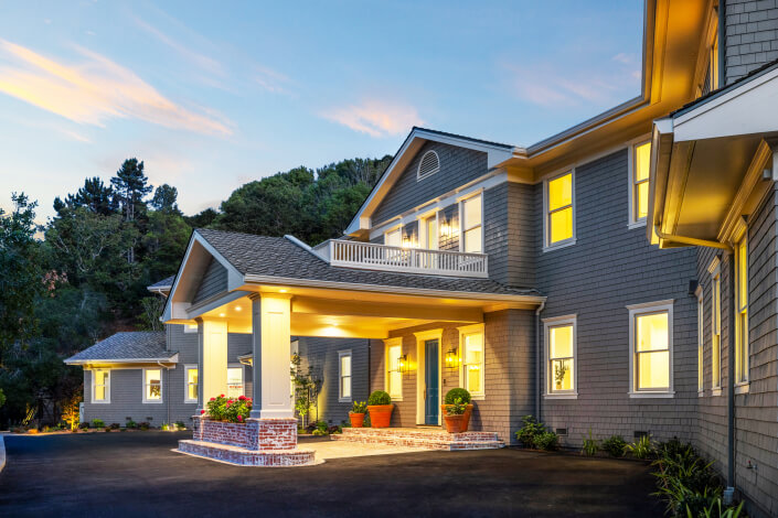 A large, two-story house at 25 Maoli Drive features grey siding and white trim, illuminated by warm lights at dusk. The entrance boasts tall columns, potted plants, and a covered porch, all surrounded by lush greenery and trees.