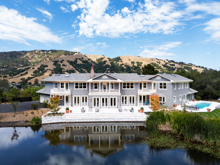 Large two-story gray house with many windows and a spacious patio, set beside a pond at 25 Maoli Drive San Rafael. The background features rolling hills, green trees, and a partly cloudy blue sky. A swimming pool sits to the right of the house.