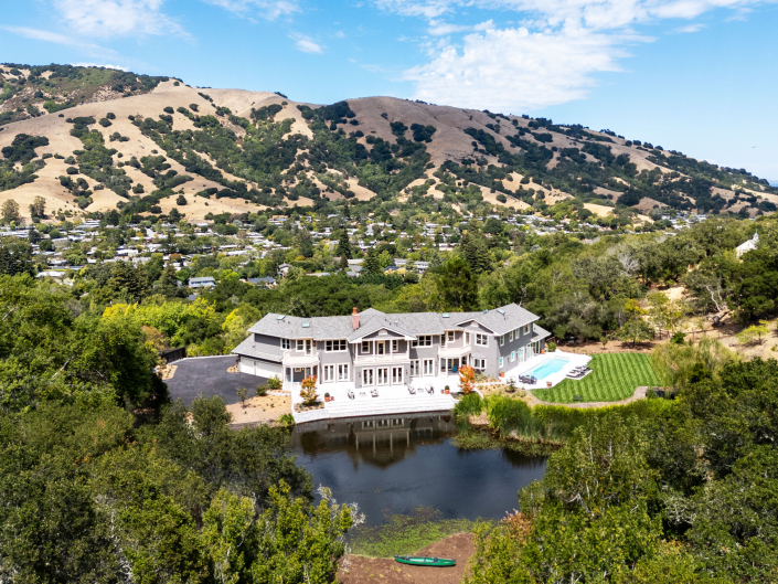 A large, modern house with a pool sits beside a pond at 25 Maoli Drive San Rafael, surrounded by trees and hills, with a small town visible in the background under a partly cloudy sky.