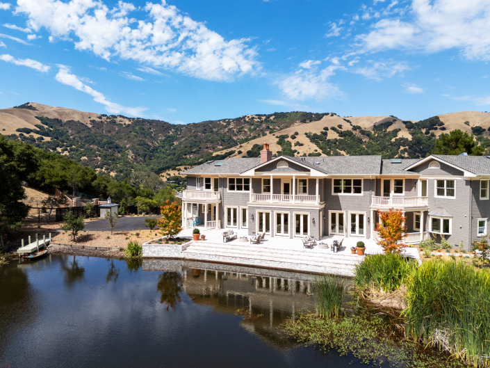 A large, modern lakeside house with a spacious patio sits beside a small pond, surrounded by hills, trees, and a blue sky with scattered clouds.