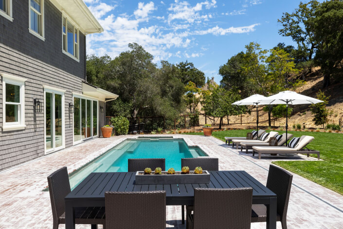 Modern backyard with a rectangular pool, surrounded by a brick patio. Black patio table and chairs in the foreground; lounge chairs and umbrellas on grass to the right. Trees and hills in the background.