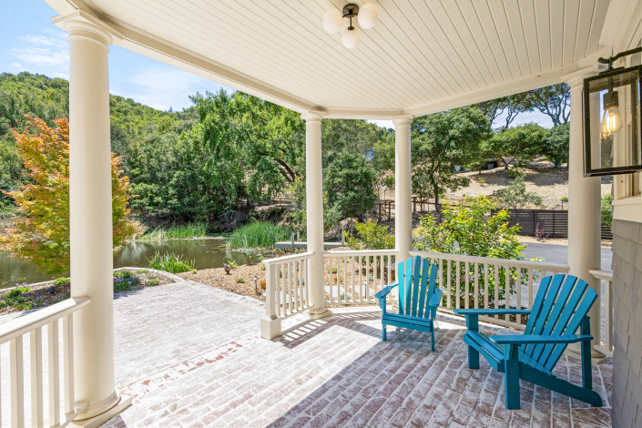 A spacious porch at 25 Maoli Drive features white columns, two blue Adirondack chairs, brick flooring, and beautiful views of a pond, trees, and a hillside under a clear sky.