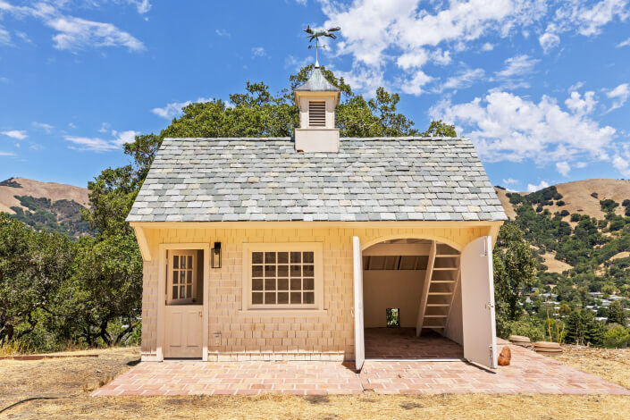 A small, light-colored cottage with a shingled roof, cupola, and weather vane sits on a brick patio at 25 Maoli Drive. One side is enclosed with a window and door; the other is open with a ladder leading to a loft. Hills and trees are in the background.