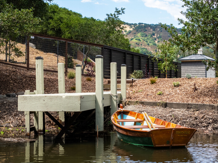 A small wooden rowboat named THISTLE is tied to a dock on a calm pond, surrounded by trees, a fence, and a shed, with hills visible in the background under a partly cloudy sky.