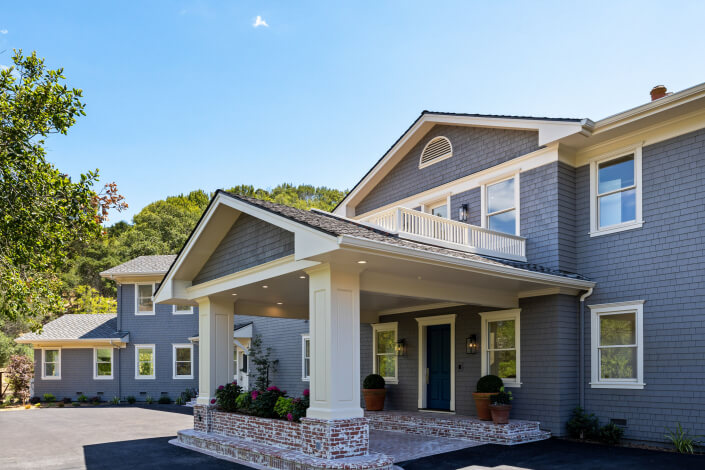 A large, two-story gray house at 25 Maoli Drive San Rafael features a covered entry supported by columns, brick steps, and white trim, set against a backdrop of trees and blue sky.
