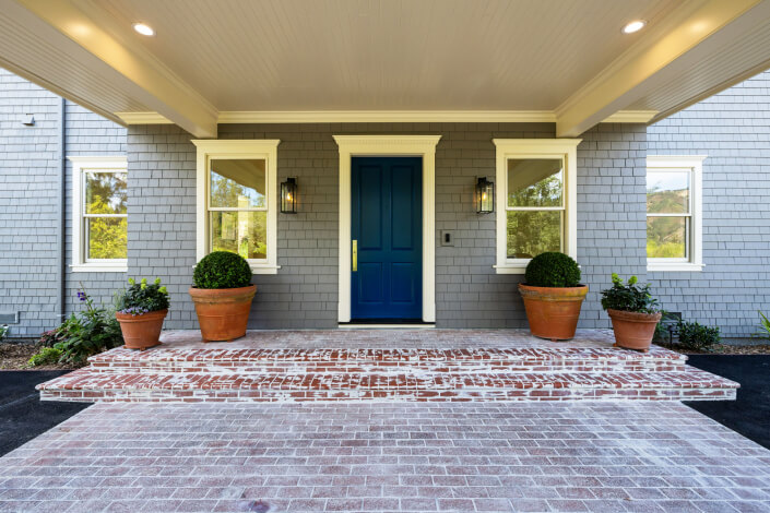 Covered front porch with brick steps, a blue door, two large potted plants on either side, two windows with cream trim, gray shingle siding, and wall-mounted lantern lights.