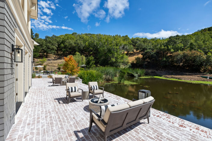 Patio with cushioned chairs and tables on a brick surface overlooks a small pond and green hills under a bright blue sky with scattered clouds.