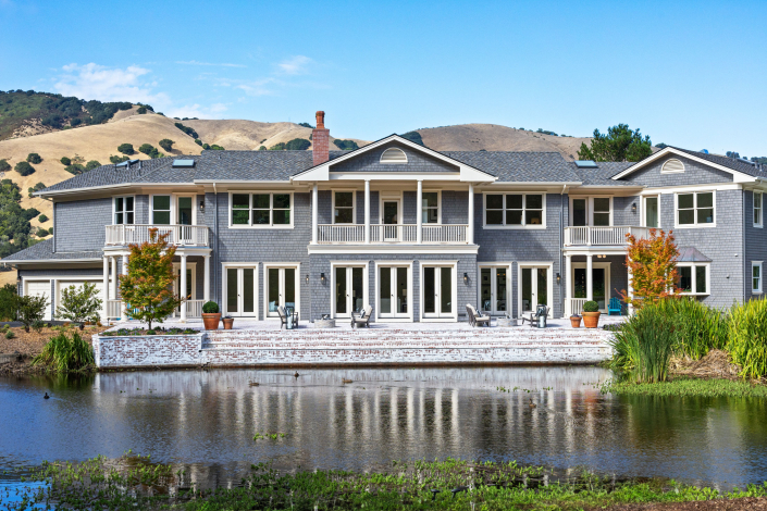 A large two-story gray house at 25 Maoli Drive San Rafael, with balconies and many windows, is reflected in a pond in the foreground, set against scenic hills and a blue sky.