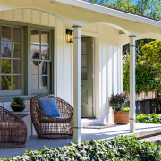 A cozy front porch at 114 Richardson Drive Mill Valley features two wicker chairs, a small table with potted plants, a white exterior, green-trimmed windows, and lush greenery surrounding the steps and garden area.