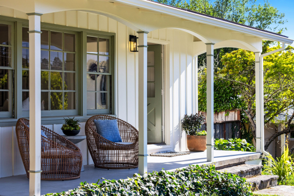 A cozy front porch at 114 Richardson Drive Mill Valley features two wicker chairs, a small table with potted plants, a white exterior, green-trimmed windows, and lush greenery surrounding the steps and garden area.