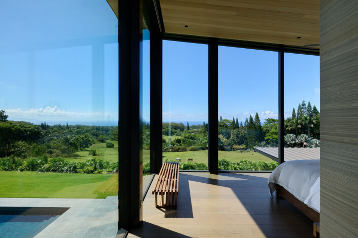 A modern bedroom with floor-to-ceiling glass windows offers a panoramic view of lush greenery, distant trees, and a clear blue sky. A wooden bench sits near the windows, and a pool is visible outside.