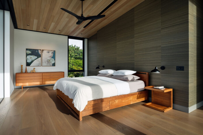 A modern Maui estate bedroom with a wooden bed frame, white bedding, nightstand, and dresser. Large windows fill the space with natural light, while a painting and decorative items accent the wood ceiling and sleek ceiling fan.