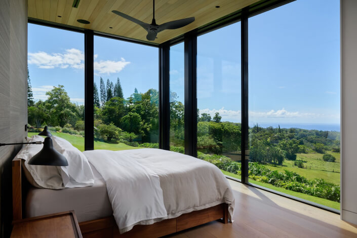 A modern bedroom in Haiku HI with large floor-to-ceiling windows offering a scenic view of green hills, trees, and blue sky. The room at 1642 W. Kuiaha Rd features a neatly made bed with white bedding and black reading lamps.