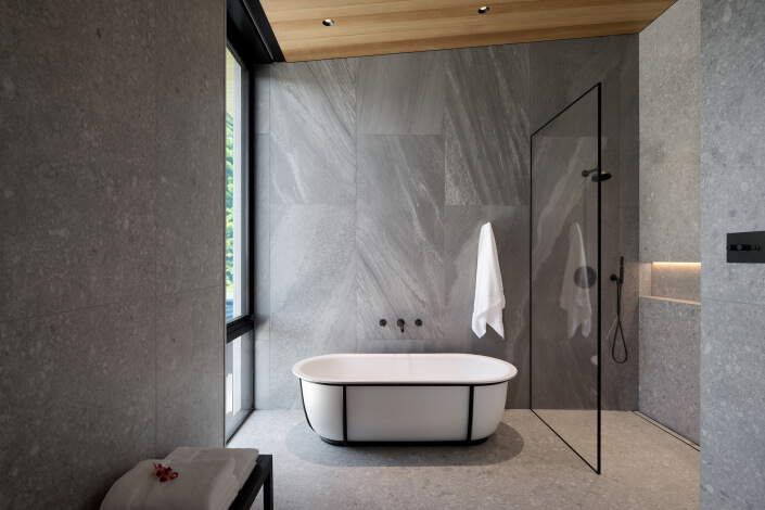 Modern bathroom with a white freestanding bathtub, gray stone walls and floor, glass shower enclosure, towel hanging on wall, and natural light from a large window. Ceiling has wood paneling. Minimalist design.