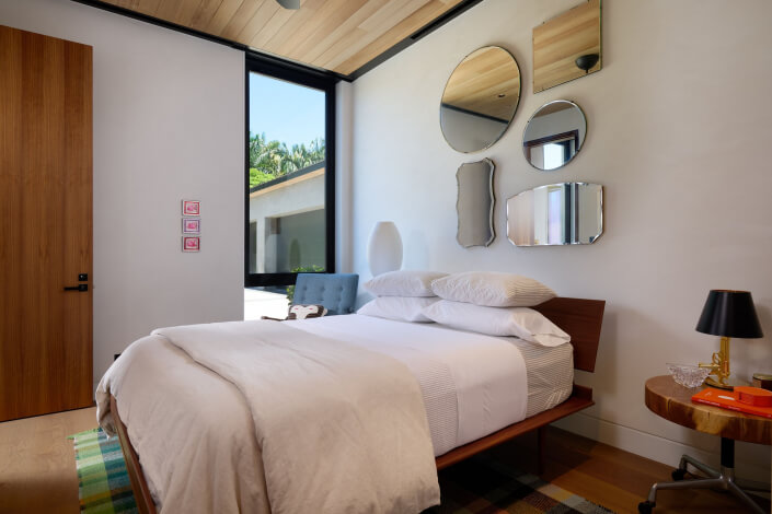 Modern bedroom with a neatly made bed, white bedding, and stacked pillows. A gallery of assorted mirrors decorates the wall above the bed. A window lets in natural light, and a wooden nightstand holds a lamp and books.