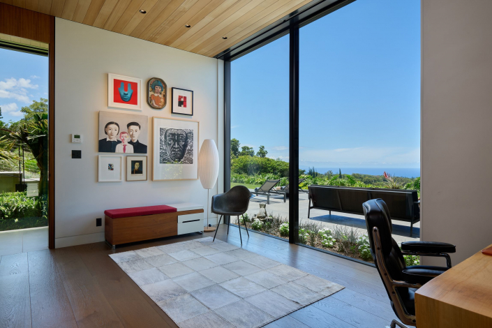 Modern home office with large floor-to-ceiling windows overlooking a garden and ocean view, contemporary artwork on the wall, a red bench, light rug, black chairs, and wooden ceiling. Bright and airy atmosphere.