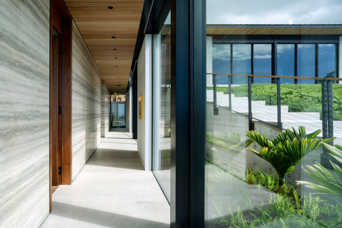 A modern hallway with floor-to-ceiling windows overlooks a lush green garden. The space features sleek concrete floors, wooden ceiling accents, and light stone walls, with natural light streaming in.