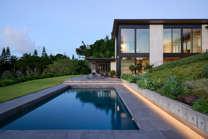 Modern house with large glass windows overlooking a rectangular swimming pool, surrounded by lush greenery and landscaped plants, with outdoor seating on a patio area at sunset.
