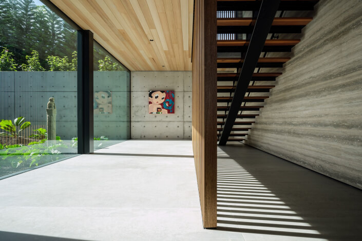 A modern Maui estate with minimalist interiors features large glass windows, concrete walls, a wooden ceiling, open stairs, and light streaming in. Two colorful art pieces hang on the wall, and lush greenery is visible outside.