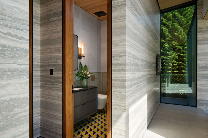 A modern bathroom with striped stone walls, a wooden door frame, patterned tile floor, a vanity with a mirror, a toilet, and a vase of flowers. Large window overlooks lush green trees outside.