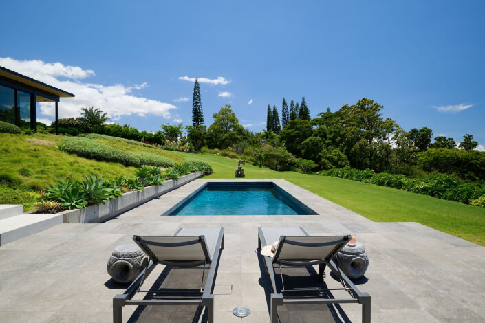 Two lounge chairs face a rectangular pool in a modern Maui estate backyard, surrounded by lush grass, plants, and trees under a clear blue sky. A glass-walled house is visible on the left.