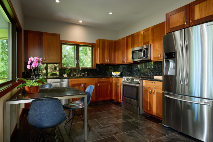 Modern kitchen with wooden cabinets, stainless steel appliances, a black stone countertop, and a small dining table. There are blue chairs and a potted orchid on the table, with large windows letting in natural light.