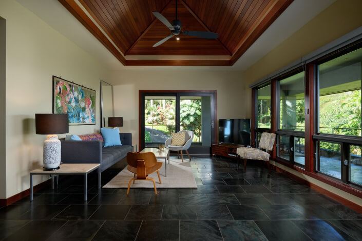 Modern living room with slate tile floor, wooden vaulted ceiling, large windows, a gray sofa, armchair, coffee table, lamps, TV, and nature views outside. Decor includes colorful artwork and neutral-toned furnishings.