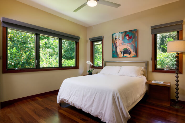 A bright bedroom with large windows, a ceiling fan, wooden floor, a bed with white bedding, a nightstand with a lamp, an orchid, and a colorful painting above the headboard. Lush greenery is visible outside.