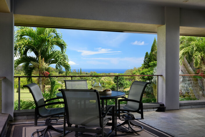 A covered patio with a glass table and four chairs overlooks a lush garden with palm trees and a distant view of the ocean under a blue sky.