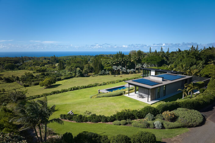 A modern house at 1642 W. Kuiaha Rd, Haiku HI features solar panels, a large manicured lawn surrounded by lush trees, a swimming pool, and ocean views—all under a clear blue sky.