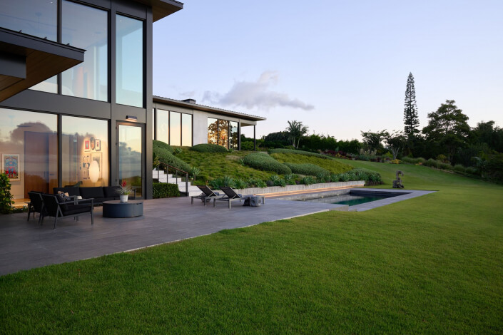 Modern house with large glass windows, patio seating, and a small rectangular pool. The surrounding area is lush green lawn and manicured bushes, with trees and a clear sky in the background.