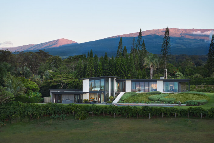 A modern Maui estate with large glass windows sits amid lush greenery, with tall pine trees and a mountain range visible in the background under a clear sky.