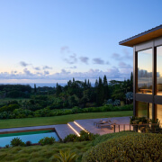 Modern Maui estate with large glass windows surrounded by lush greenery, landscaped garden, and a swimming pool, set against a backdrop of tall trees under a clear evening sky.