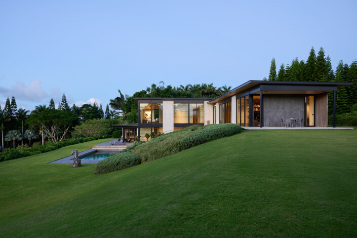 Modern Maui estate with large windows sits on a grassy hill, featuring a patio, swimming pool, and surrounded by lush greenery and tall trees under a clear blue sky.