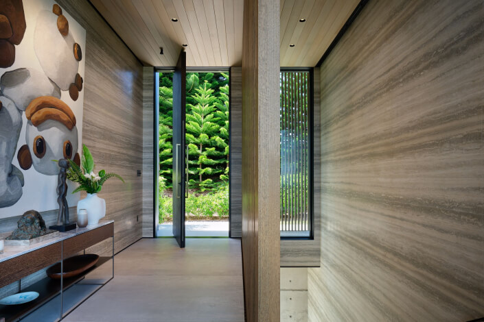 A modern hallway with light stone walls, a console table decorated with vases and art, large abstract wall art, and a tall glass door revealing lush greenery outside. Vertical windows bring in natural light.