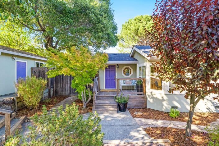 A charming house at 102 Clorinda Avenue in Gerstle Park, San Rafael, features a light gray exterior, a purple front door, white porch bench, wooden steps, potted plants, and trees under a clear blue sky.