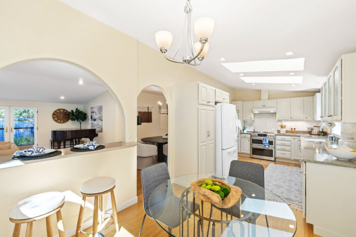 Bright, open kitchen and dining area with white cabinets, modern appliances, a glass table with gray chairs, and a bowl of green apples. Arched openings connect to a living room with a piano and large clock.