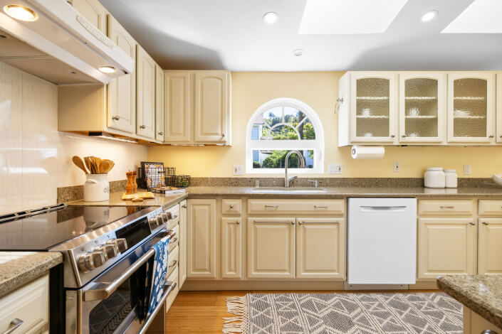 Bright kitchen with cream cabinets, stainless steel oven, dishwasher, and sink under a round window. Countertops are stone, shelves hold dishes, and a patterned rug covers the wooden floor. Sunlight fills the room.