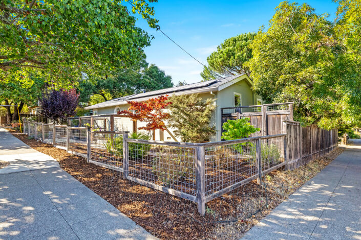 Single-story house with a light exterior, surrounded by a wooden and wire fence. Lush trees and plants fill the yard. The home is located on a corner lot next to sidewalks and driveways, under a clear blue sky.