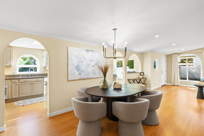 A modern dining room at 102 Clorinda Avenue, Gerstle Park, San Rafael features a round black table, four light gray chairs, wood flooring, a contemporary chandelier, and arched doorways to a bright kitchen and living area.