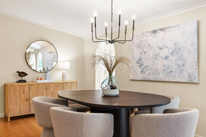 Modern dining room with a round black table, four beige chairs, a glass vase with dried grass centerpiece, light wood sideboard, round mirror, abstract wall art, and a black chandelier.