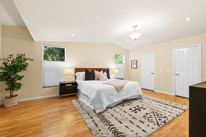 Bright bedroom with a large bed, nightstands, lamps, and neutral decor. Hardwood floor, abstract art, black bench, potted plant, and two windows add to the airy, inviting atmosphere.