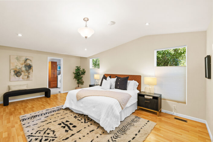 Bright bedroom with a large bed, nightstands, lamps, and neutral decor. Hardwood floor, abstract art, black bench, potted plant, and two windows add to the airy, inviting atmosphere.