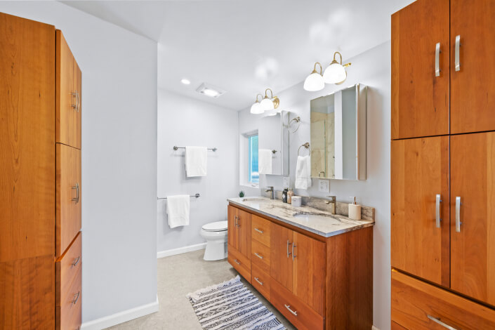 Modern bathroom at 102 Clorinda Avenue Gerstle Park San Rafael features wooden cabinets, a double-sink marble vanity, wall-mounted mirror, glass fixtures, towel racks with white towels, a striped rug, and a toilet by the window.