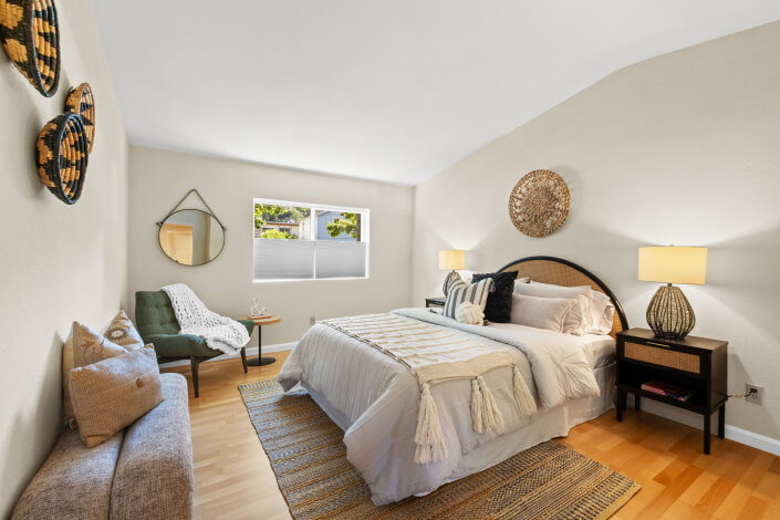 A cozy bedroom with a neatly made bed, two nightstands with lamps, a large mirrored closet, light wood floor, a blue chair, and soft neutral decor. A hallway and bathroom are visible through an open door.