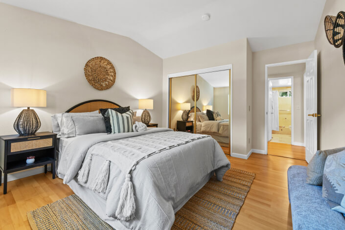 A cozy bedroom with a neatly made bed, two nightstands with lamps, a large mirrored closet, light wood floor, a blue chair, and soft neutral decor. A hallway and bathroom are visible through an open door.