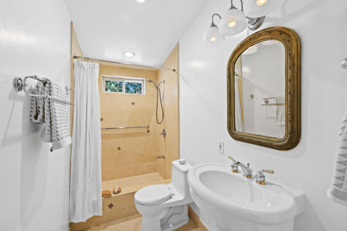 A bright bathroom with a walk-in shower, beige tiles, a white shower curtain, a toilet, a pedestal sink, a large ornate mirror, and white walls. Towels hang on racks, and the shower has a grab bar and window.